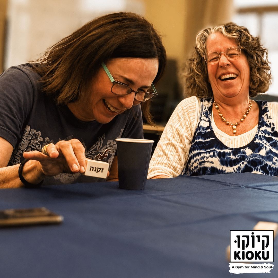 שתי משתתפות צוחקות סביב שולחן, אחת מחזיקה אבן עץ עם המילה זקנה